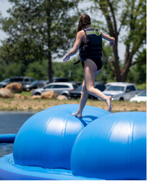 A person in a life jacket balances on a large blue inflatable structure on a lake, with cars and trees in the background.
