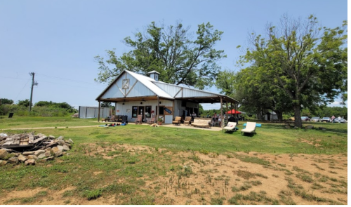 A rustic building with a metal roof, surrounded by green grass and trees, featuring outdoor seating and a fire pit.