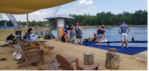 A group of children in swim gear gather by a lake, with a watersports tower and picnic tables nearby.