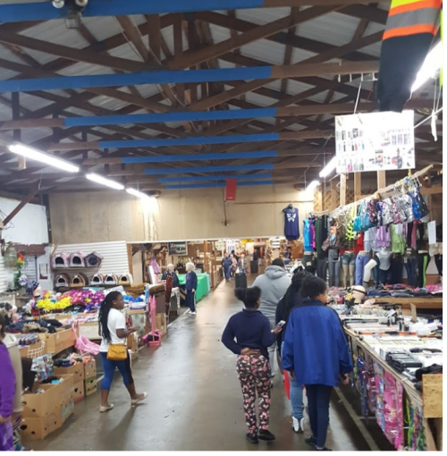 A bustling market interior with shoppers browsing stalls filled with clothing and accessories under a wooden ceiling.