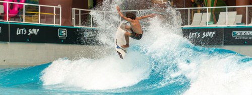 A surfer performs a trick on a wave in a surf park, with splashes of water around him.