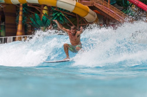 A man surfing on a wave in a water park, with colorful water slides in the background.
