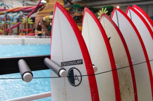 A row of surfboards with red accents lined up near a pool, with a water park in the background.