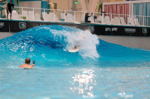 A surfer rides a large artificial wave while another person captures the moment with a camera.