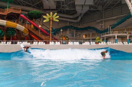 Two surfers ride a wave in an indoor water park, surrounded by slides and tropical decor.