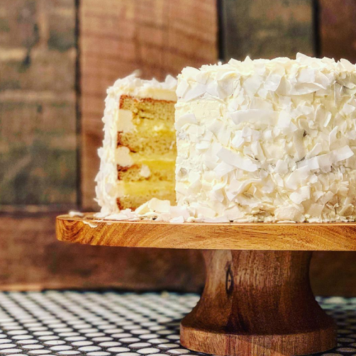 A layered coconut cake on a wooden cake stand, with shredded coconut on top and a slice cut out.