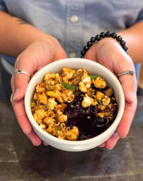 A person holds a bowl of popcorn topped with colorful toppings, set against a blurred background.