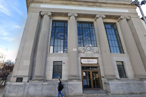 Historic building with large columns, a clock, and glass windows; a person walks by in front.