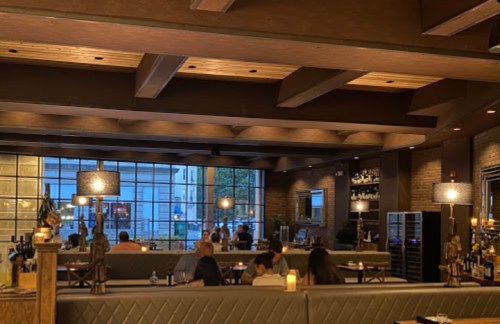 Cozy restaurant interior with dim lighting, wooden beams, and patrons dining at tables near large windows.