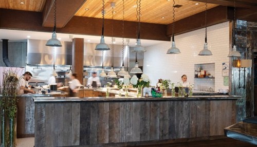 A modern restaurant kitchen with chefs working, featuring a wooden counter and industrial lighting.