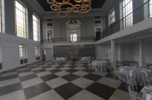 A spacious event hall with a checkered floor, elegant tables, and large windows, featuring a modern chandelier.