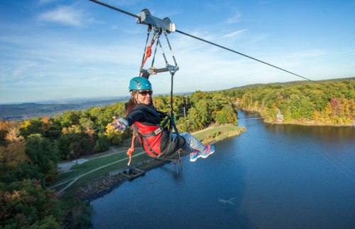 A person zip-lining over a scenic landscape with trees and a lake, wearing a helmet and smiling against a clear blue sky.