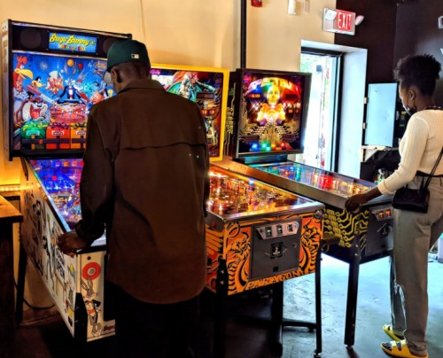 Two people playing pinball machines in a vibrant arcade setting, with colorful game graphics visible.