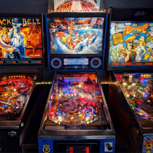 A colorful pinball machine titled "River Poster" surrounded by other vintage pinball games in a lively arcade setting.