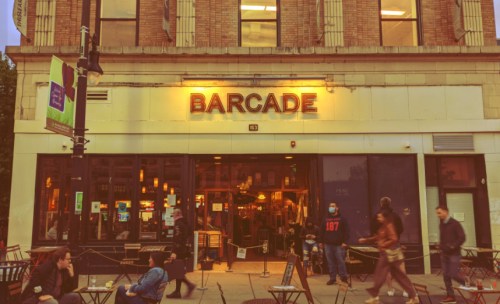 A lively street scene in front of a barcade, with people walking and sitting outside as evening lights glow.