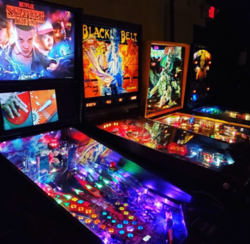 A vibrant arcade featuring pinball machines, including "Stranger Things" and "Black Belt," illuminated with colorful lights.
