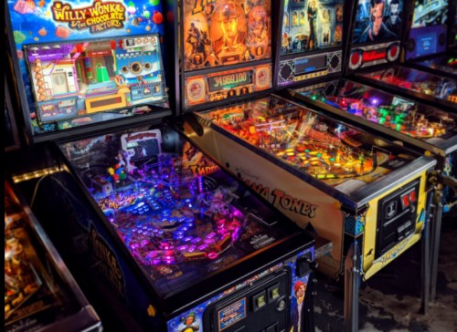 A row of colorful pinball machines, including "Willy Wonka" and "Indiana Jones," illuminated with vibrant lights.