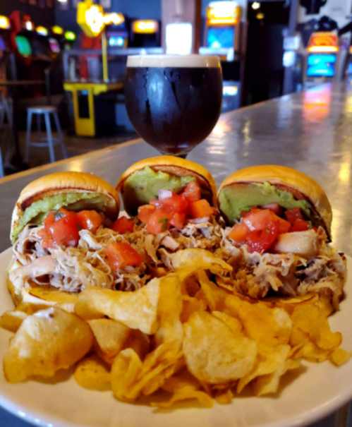 Three sliders with pulled pork, tomatoes, and guacamole, served with a side of potato chips and a dark beer in the background.