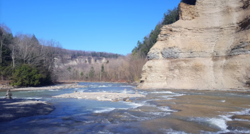 A river flows between steep, rocky cliffs under a clear blue sky, surrounded by trees and a serene landscape.