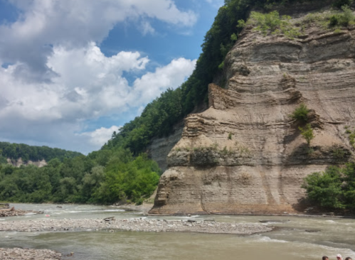 A river flows past steep, layered cliffs under a partly cloudy sky, surrounded by lush green trees.