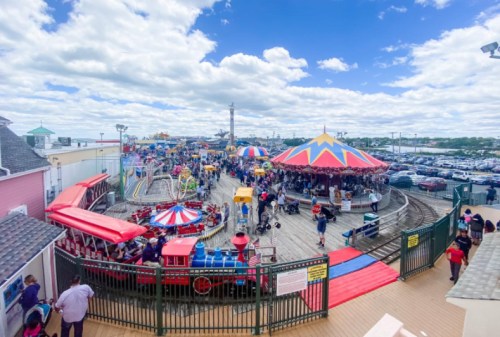A vibrant amusement park scene with a carousel, rides, and a crowd under a blue sky with fluffy clouds.