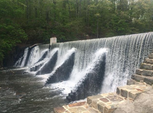 A cascading waterfall flows over a stone dam, surrounded by lush green trees and a rocky path.