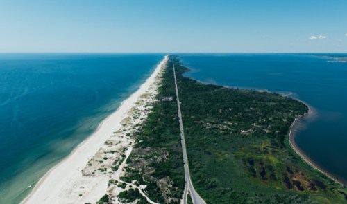 Aerial view of a long, narrow strip of land separating the ocean from a lush green area, with a road running through it.