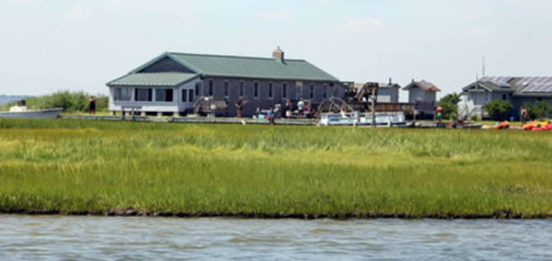 A gray building near a marshy area, surrounded by green grass and water, with boats visible in the background.