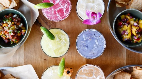 A vibrant spread of colorful cocktails and fresh salsa served with tortilla chips on a wooden table.