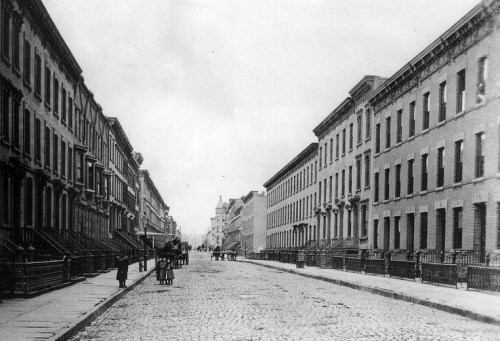 Black and white photo of a cobblestone street lined with brick buildings and people walking in the distance.