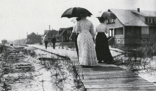 Two women in long dresses walk along a boardwalk with umbrellas, while others stroll in the background near beach houses.
