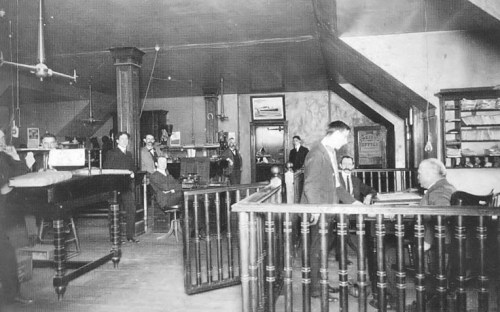 Historic interior of a busy office with men engaged in conversation and work, featuring wooden furniture and a staircase.