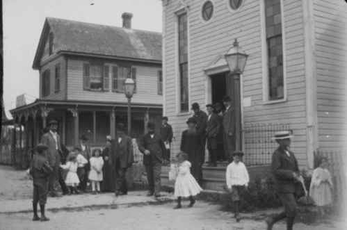 A historic black-and-white photo of people in formal attire outside a building, with children playing nearby.