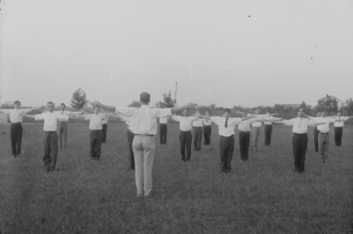A group of men in white shirts and ties standing in a field, arms outstretched, following a leader in front.
