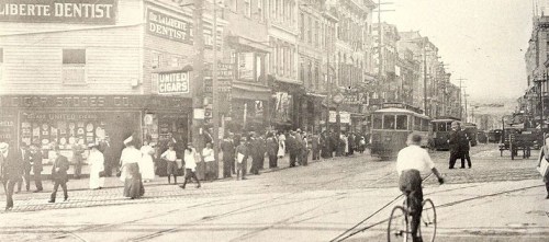 Historic street scene with pedestrians, shops, and a streetcar, capturing early 20th-century urban life.