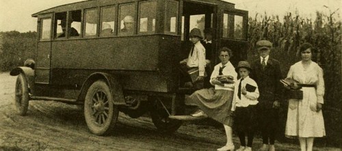 A vintage bus parked on a dirt road with a group of people, including children, standing beside it.