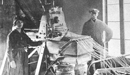 A historical black-and-white photo of a woman operating machinery, with a man standing beside her in a factory setting.