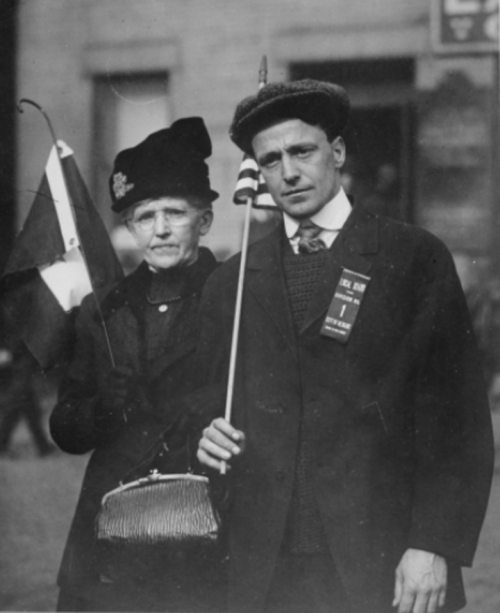 A man and woman stand together, holding flags and wearing vintage clothing, in a historical black-and-white photo.