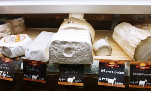 A display of various cheeses with labels, including Sofia, Leonora, and Monte Enebro, in a cheese shop.