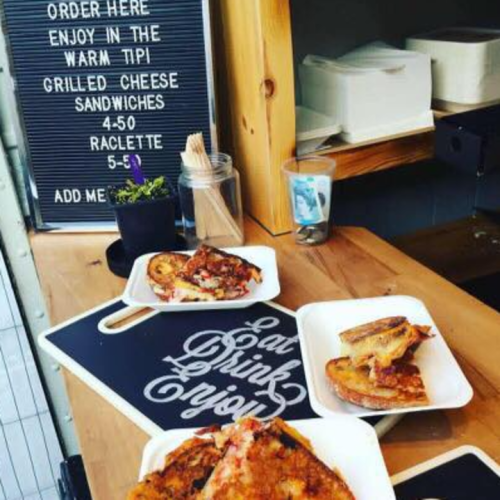 A cozy food stall with a menu board, featuring grilled cheese sandwiches and raclette, alongside a plate of food.