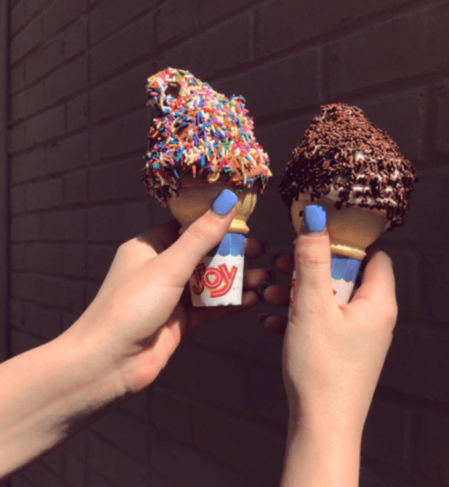 Two hands holding ice cream cones with colorful sprinkles and chocolate sprinkles against a brick wall.
