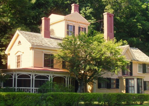 A charming yellow Victorian house with a wraparound porch, surrounded by lush greenery and tall trees.