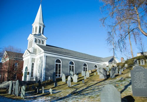 A gray church with a tall steeple beside a sloping graveyard under a clear blue sky. Snow lightly covers the ground.