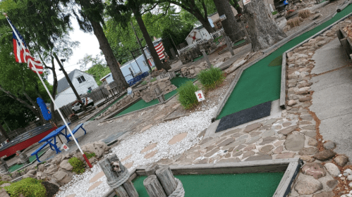 A mini-golf course with green turf, stone pathways, and trees, featuring American flags in the background.