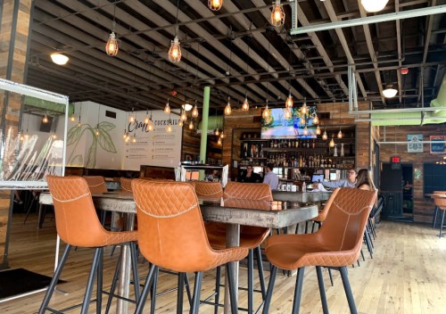 A modern restaurant interior with wooden decor, bar seating, and pendant lights, featuring a bar and TV in the background.