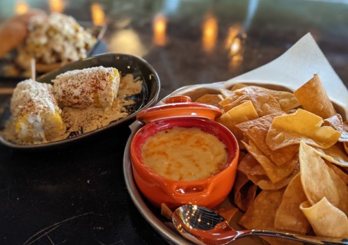 A plate of tortilla chips with cheese dip, alongside elote topped with cheese and spices.