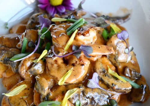 A close-up of shrimp pasta topped with creamy sauce, edible flowers, and green onions on a white plate.