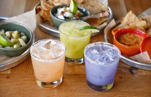 Three colorful cocktails sit on a wooden table, surrounded by chips and dips in bowls.