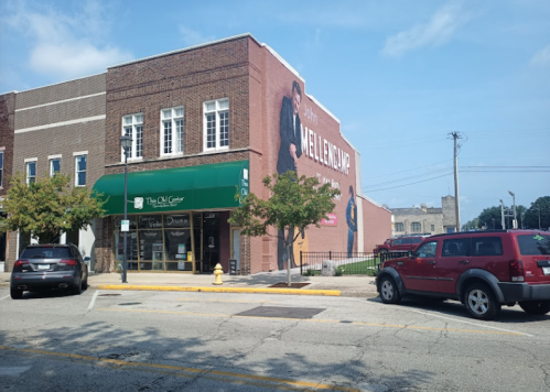 A brick building with a green awning and a large mural on the side, parked cars on the street, and trees nearby.