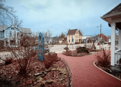 A quaint town square with brick pathways, surrounded by trees and historic buildings under a cloudy sky.